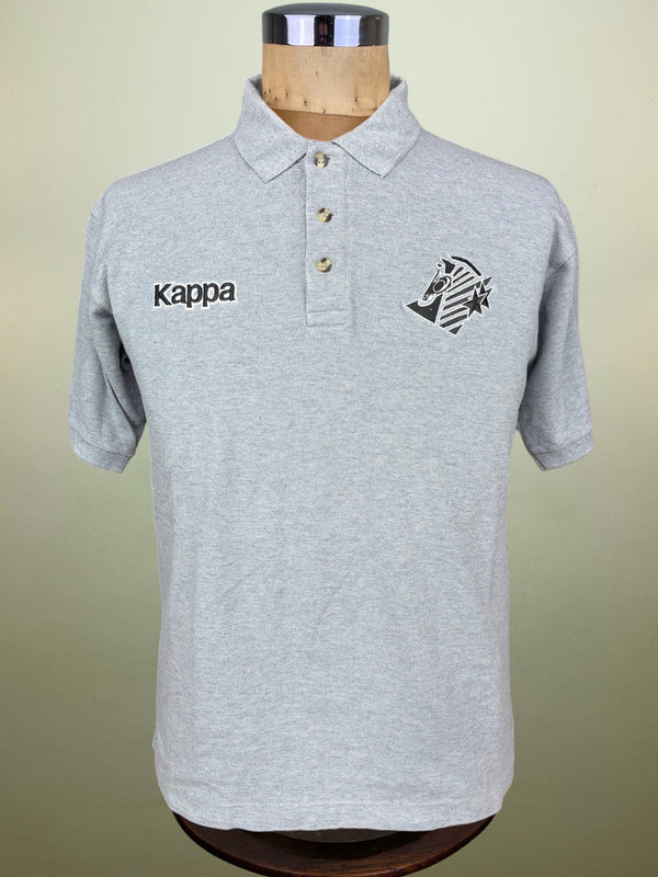 A light gray heathered Kappa polo shirt displayed on a wooden dress form mannequin against a soft beige background. The shirt features the black Kappa logo embroidered on the right chest and the club crest on the left chest.
