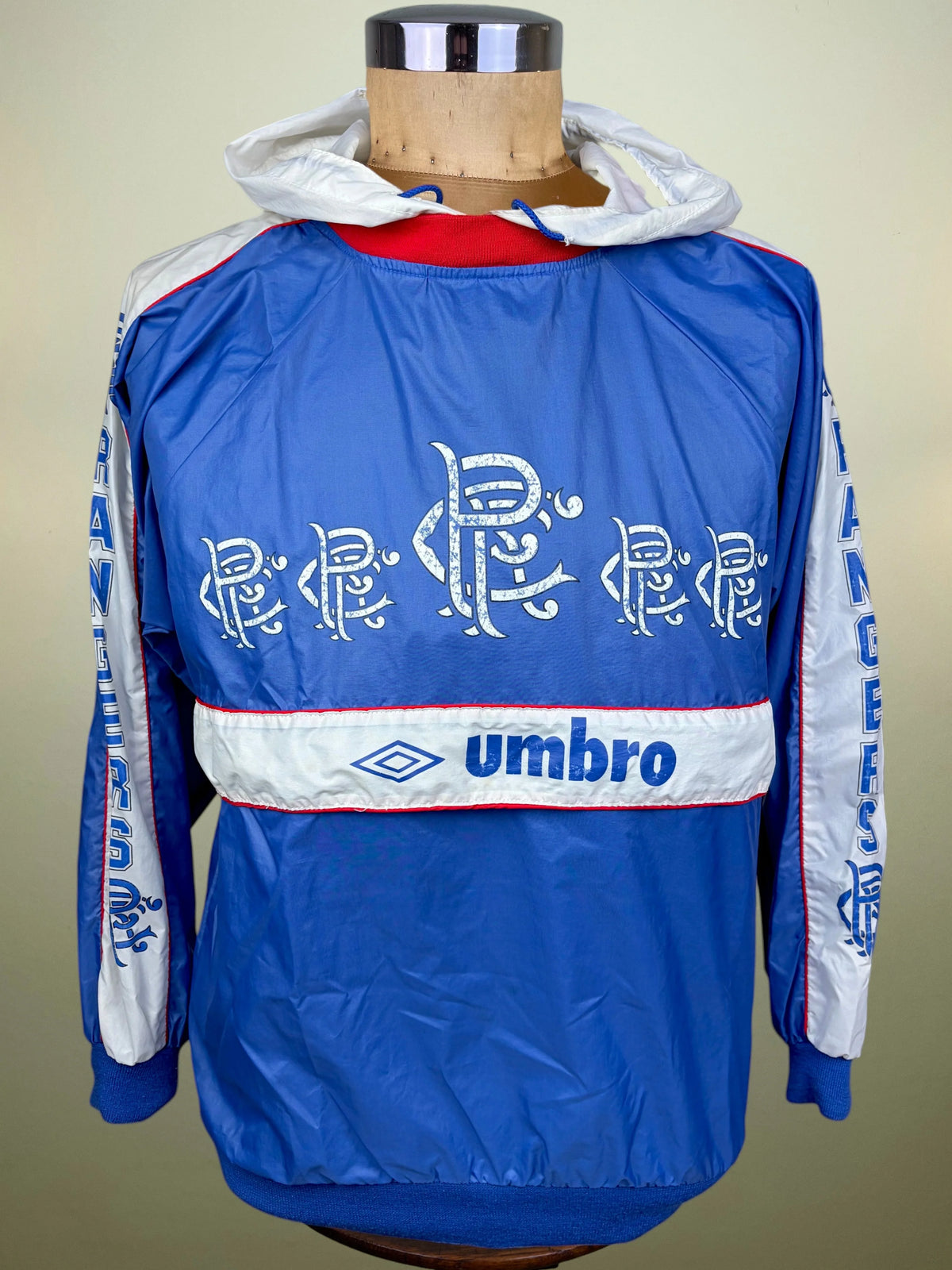 A blue Umbro windbreaker jacket displayed on a mannequin torso. The jacket features a white horizontal stripe across the chest with the blue Umbro logo