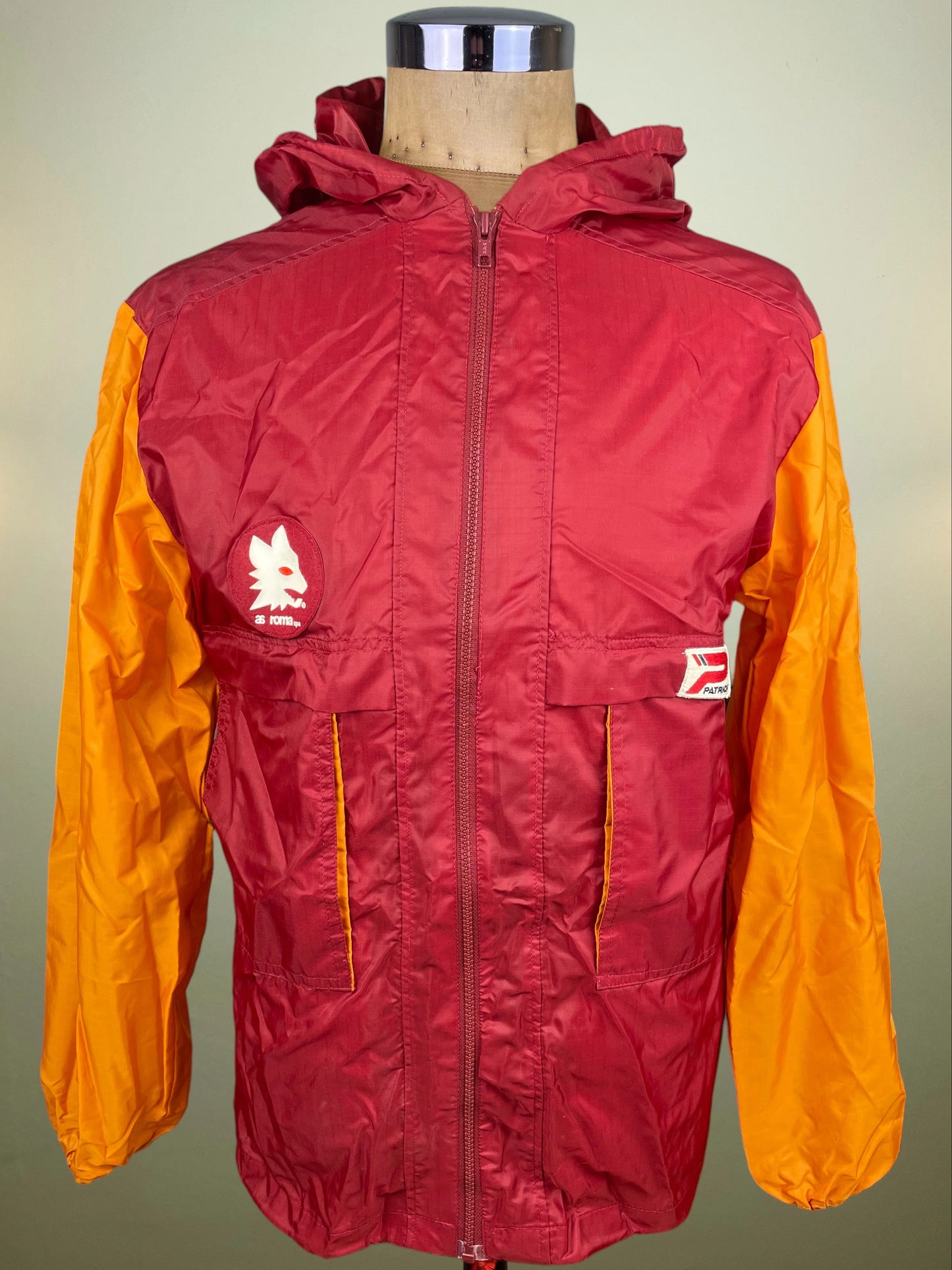 A hooded zip-front AS Roma jacket with lupetto crest on the chest and Patrick logo displayed on a mannequin torso against a plain light background. The jacket has a dark red body with bright orange sleeves and side panels