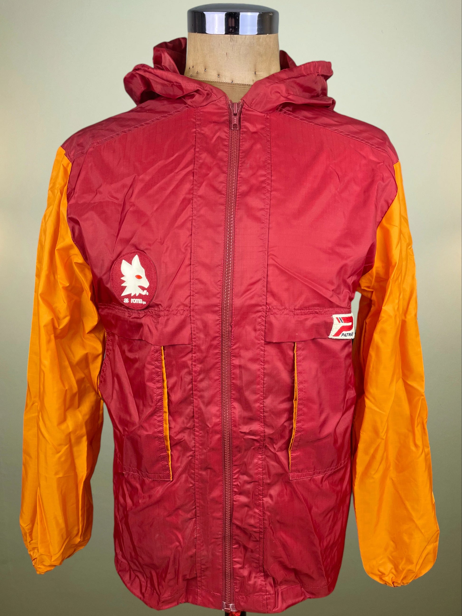 A hooded zip-front AS Roma jacket with lupetto crest on the chest and Patrick logo displayed on a mannequin torso against a plain light background. The jacket has a dark red body with bright orange sleeves and side panels