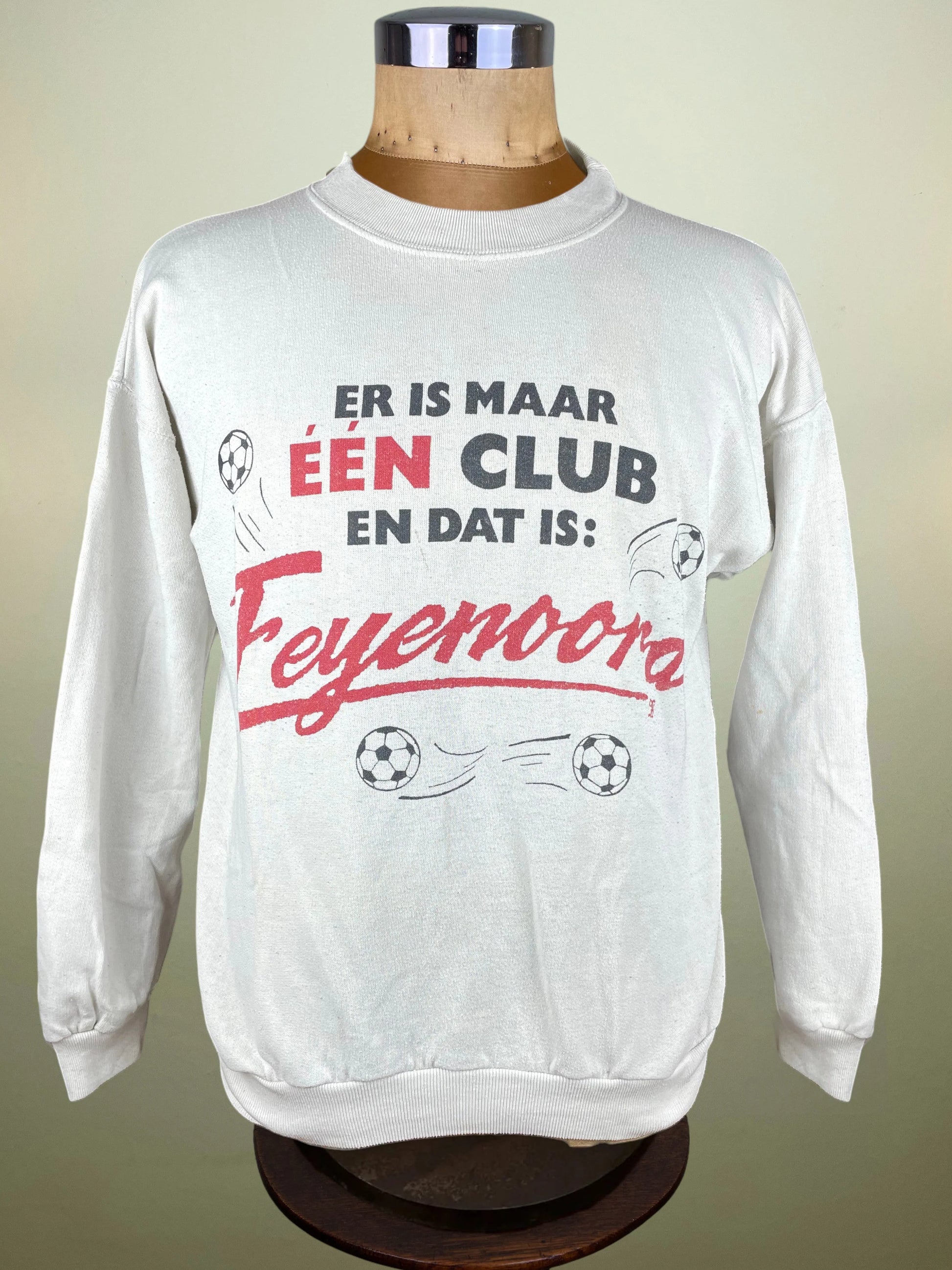 A white long-sleeve sweatshirt displayed on a wooden dress form mannequin. The sweatshirt features a graphic print in Dutch with the text 'ER IS MAAR ÉEN DAT IS: Feyenoora' in red and black lettering