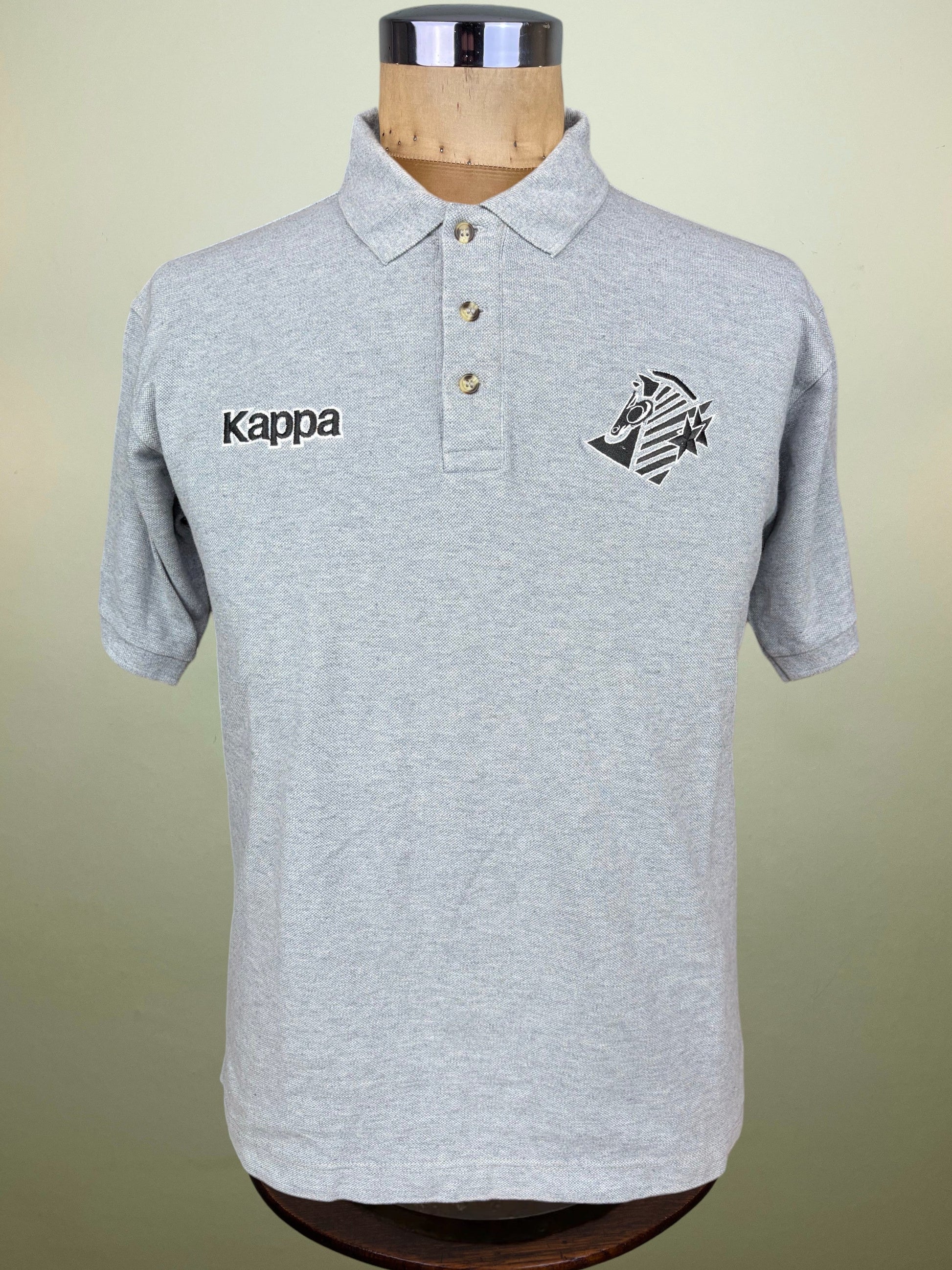 A light gray heathered Kappa polo shirt displayed on a wooden dress form mannequin against a soft beige background. The shirt features the black Kappa logo embroidered on the right chest and the club crest on the left chest.