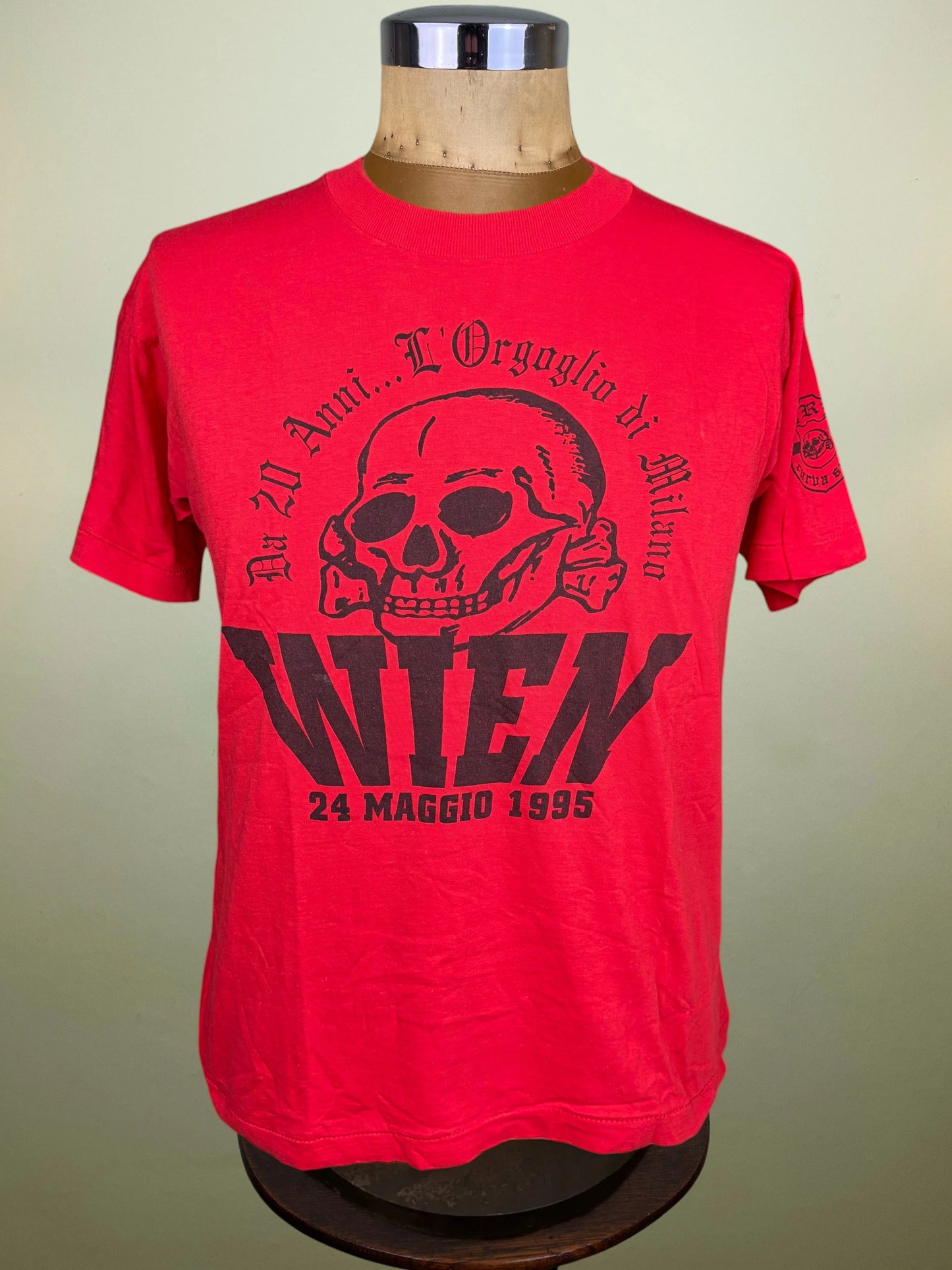 A bright red short-sleeve t-shirt displayed on a wooden mannequin torso against a light beige background. The shirt features a black graphic print of a skull and gothic text reading 'Da 20 Anni... L'Orgoglio di Milano" and then in bold "Wien 24 Maggio 1995"